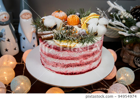 Christmas cake made of chocolate base with cream, decorated with berries and fruits on the background of candles, garlands, fir branches and cones 94954974