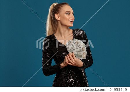 Blonde model with ponytail, in black sequin dress. She is smiling, holding some cash, posing on blue background. Gambling, poker, casino. Close-up Blonde model with ponytail, in black sequin dress. She is smiling, holding some cash, posing on blue background. Gambling, poker, casino. Close-up 94955349
