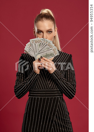Blonde lady with ponytail, in black dress in rhinestones. Showing fan of hundred dollar bills, posing on red background. Poker, casino. Close-up Blonde lady with ponytail, in black dress in rhinestones. Showing fan of hundred dollar bills, posing on red background. Poker, casino. Close-up 94955354