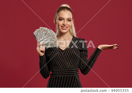 Blonde model with ponytail, in black dress in rhinestones. She is smiling, holding some cash, posing on red background. Poker, casino. Close-up 94955355