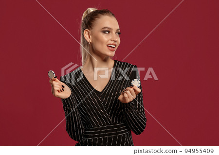 Blonde female with ponytail, in black dress in rhinestones. She smiling, showing two white chips, posing on red background. Poker, casino. Close-up Blonde female with ponytail, in black dress in rhinestones. She smiling, showing two white chips, posing on red background. Poker, casino. Close-up 94955409