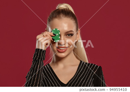 Blonde girl with ponytail, in black dress in rhinestones. She is smiling, showing two green chips, posing on red background. Poker, casino. Close-up 94955410