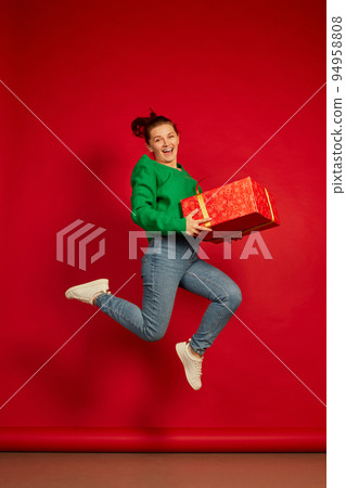 Portrait of happy smiling woman in green sweater jumping in joy with present box isolated over red studio background 94958808