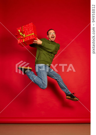Portrait of young cheerful man jumping in joy with present box isolated over red studio background. Super excited 94958812