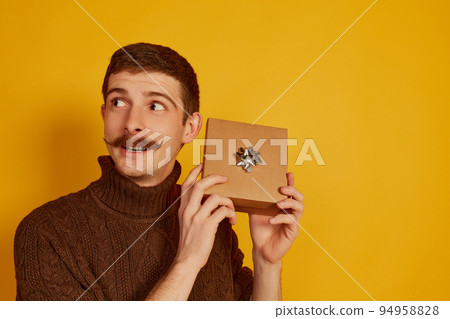 Portrait of young man in brown sweater holding little present box with bow decoration over yellow background. Portrait of young man in brown sweater holding little present box with bow decoration over yellow background. 94958828