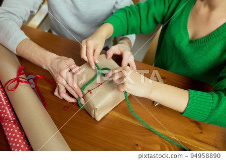Top view image of man and woman wrapping presents together at home, using wrapping paper and ribbons. Making bow with string Top view image of man and woman wrapping presents together at home, using wrapping paper and ribbons. Making bow with string 94958890