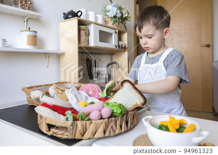 Cute little baby boy in chef hat and apron playing at childish kitchen cooking food back view Cute little baby boy in chef hat and apron playing at childish kitchen cooking food back view 94961323