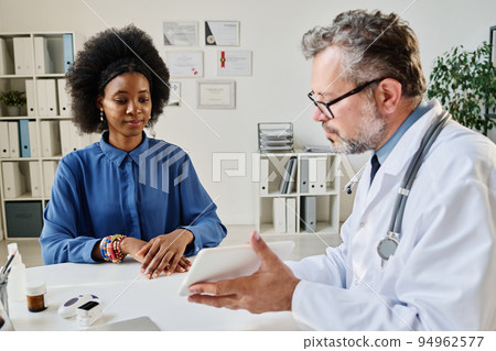 Mature doctor showing the results of analysis on digital tablet to patient during her consultation at office Mature doctor showing the results of analysis on digital tablet to patient during her consultation at office 94962577