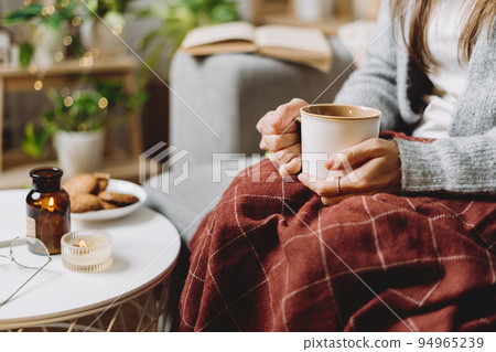 Cozy woman legs in knitted winter warm sweater and checkered plaid drinking hot cocoa or coffee in mug, sitting on couch at home. Autumn vibes with candle, decor, cookies and indoor plants. 94965239