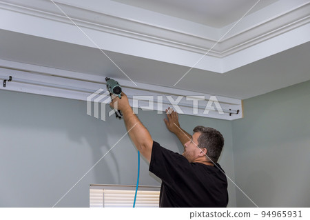 There is a professional handyman who is nailing wooden cornices on the curtain wall over the window of a newly constructed home. 94965931