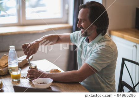 Man in the kitchen having breakfast and putting chocolate paste on bread Man in the kitchen having breakfast and putting chocolate paste on bread 94966296