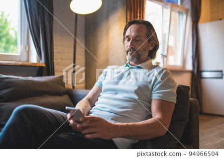 Bearded man sitting in the armchair awith a smartphone in hand Bearded man sitting in the armchair awith a smartphone in hand 94966504