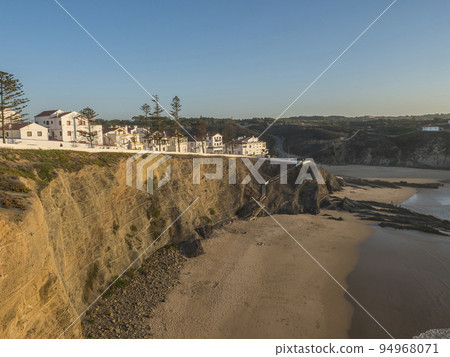 View of white houses of Zambujeira do Mar from the sand beach Praia da Zambujeira do Mar,sea shore with cliffs and rock, sunny day, clear blue sky. Rota Vicentina coast, Portugal 94968071