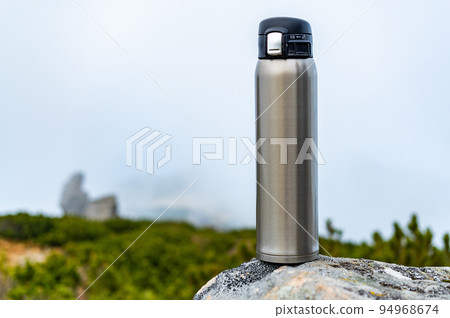 Steel thermos on a stone. Steel bottle on a stone in the mountains. Bottle on the background of clouds and mountains. 94968674