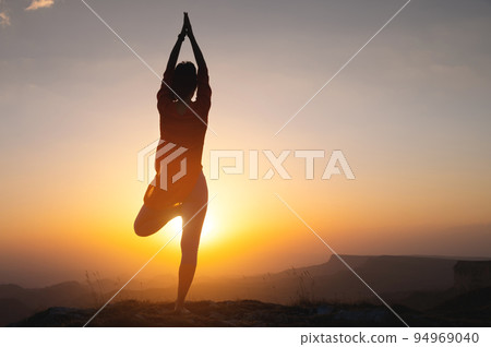 Silhouette of a young woman in a tree pose outdoors in the mountains watching the sunset, yoga balance near the cliff Silhouette of a young woman in a tree pose outdoors in the mountains watching the sunset, yoga balance near the cliff 94969040