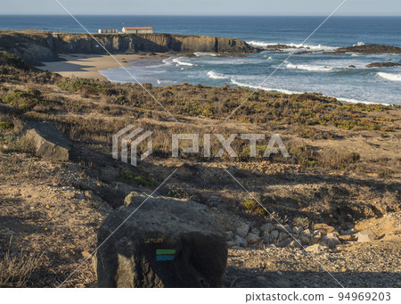 Stone with blue and green sign of Fishermans trail and view of Praia Grande de Almograve sand beach with ocean waves and small beach house. clear blue sky. Rota Vicentina coast, Almograve, Portugal 94969203