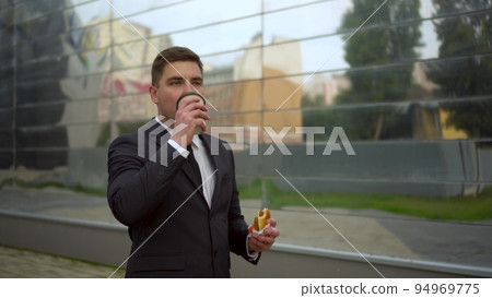 Young businessman eats fast food. A man in a suit walks next to the business center drinking coffee and eating cheeseburger. 94969775