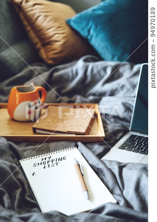 Shopping List in note book near laptop and tea coffee cup at bad in home. Shopping Preparation Consumer Buying 94970189