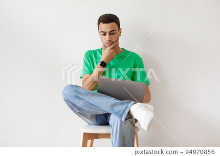 Thoughtful arab guy using laptop while sitting on chair against white wall, working or studying remotely on portable pc 94970856