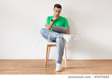 Pensive arab man using laptop and thinking, browsing internet while sitting on chair against white wall at home Pensive arab man using laptop and thinking, browsing internet while sitting on chair against white wall at home 94970888