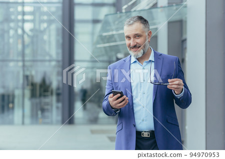 Portrait senior handsome gray-haired male teacher, professor, lecturer stands in a suit in a modern campus, holds a phone and glasses. He looks at the camera, smiles. Portrait senior handsome gray-haired male teacher, professor, lecturer stands in a suit in a modern campus, holds a phone and glasses. He looks at the camera, smiles. 94970953