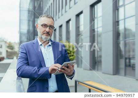 A handsome senior gray-haired man in a suit and glasses holds a tablet in his hands and reads the news. Standing near a modern office center, looking at the camera. A handsome senior gray-haired man in a suit and glasses holds a tablet in his hands and reads the news. Standing near a modern office center, looking at the camera. 94970957