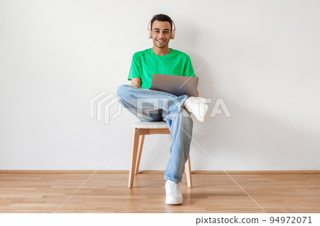 Happy middle eastern man using laptop and wearing wireless headphones, sitting on chair over white wall Happy middle eastern man using laptop and wearing wireless headphones, sitting on chair over white wall 94972071