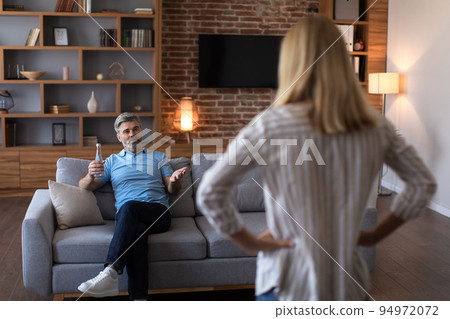 Angry mature caucasian wife scolds at husband with bottle of beer on sofa in living room interior Angry mature caucasian wife scolds at husband with bottle of beer on sofa in living room interior 94972072