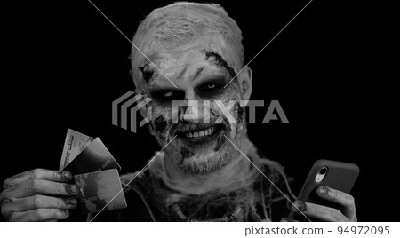 Sinister man Halloween zombie using credit bank cards and smartphone while purchases online shopping Sinister man Halloween zombie using credit bank cards and smartphone while purchases online shopping 94972095