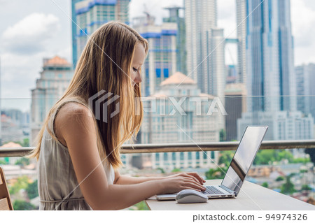 Young woman is working on a laptop on her balcony overlooking the skyscrapers. Freelancer, remote work, work from home 94974326
