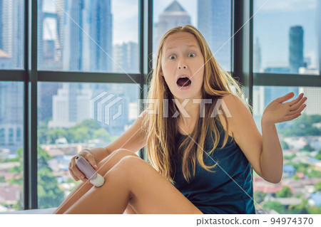 Young woman sitin on bed at home and doing epilation with epilator on legs and is in pain. On the background of a window overlooking the big city, skyscrapers Young woman sitin on bed at home and doing epilation with epilator on legs and is in pain. On the background of a window overlooking the big city, skyscrapers 94974370