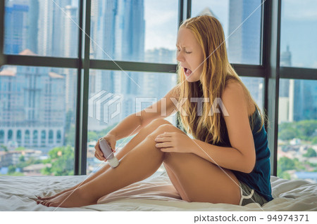 Young woman sitin on bed at home and doing epilation with epilator on legs and is in pain. On the background of a window overlooking the big city, skyscrapers Young woman sitin on bed at home and doing epilation with epilator on legs and is in pain. On the background of a window overlooking the big city, skyscrapers 94974371