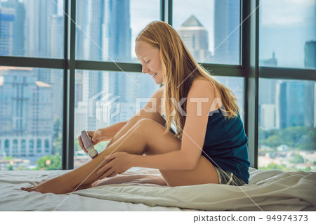Young woman sitin on bed at home and doing epilation with epilator on legs. On the background of a window overlooking the big city, skyscrapers 94974373