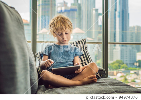 Boy uses a tablet at home on the couch in the background of a window with skyscrapers. Modern children in the megalopolis use a tablet concept Boy uses a tablet at home on the couch in the background of a window with skyscrapers. Modern children in the megalopolis use a tablet concept 94974376