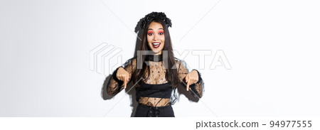 Happy beautiful asian woman in halloween gothic dress, pointing fingers down as showing announcement, demonstrating your promo, standing over white background 94977555