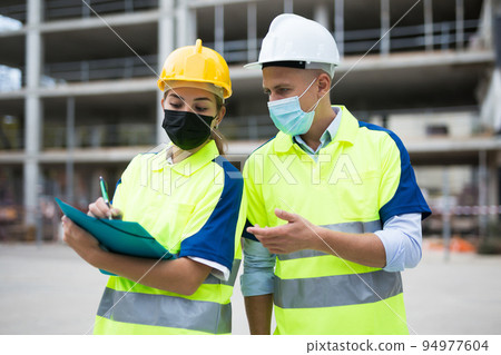 Two engineers in protective masks are discussing a construction plan Two engineers in protective masks are discussing a construction plan 94977604