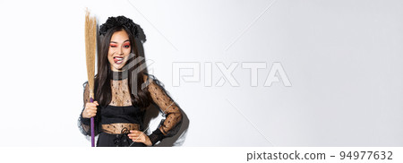 Sassy good-looking asian woman in witch costume showing tongue, holding broom and posing over white background 94977632