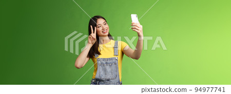 Tender cute asian brunette female, hold smartphone, posing selfie, look telephone screen, photographing, show victory peace sign, joyfully chatting friend abroad, stand green background 94977741