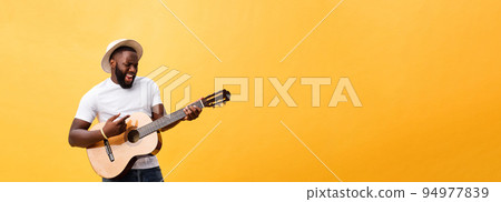 Muscular black man playing guitar, wearing jeans and white tank-top. Isolate over yellow background. Muscular black man playing guitar, wearing jeans and white tank-top. Isolate over yellow background. 94977839