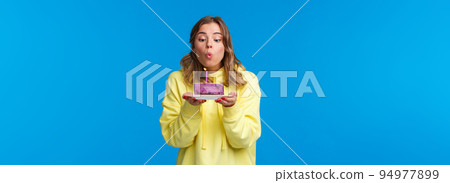 Celebration, party and lifestyle concept. Silly cute european blond woman in yellow hoodie, making wish holding fruit cake blowing out b-day candle, standing blue background at birthday party 94977899