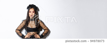 Angry and pissed-off asian female magician, evil witch in black dress and wreath looking mad at someone, frowning and looking judgemental, standing over white background 94978073