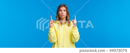 Jealous and gloomy cute blond female student feel regret not having something she desires, sobbing frowning sad as looking pointing up, standing over blue background 94978079