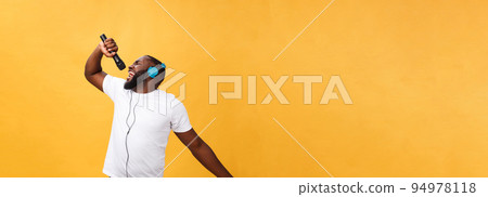 Portrait of cheerful positive chic handsome african man holding microphone and having headphones on head listening music singing song enjoying weekend vacation isolated on yellow background 94978118
