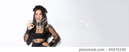 Attractive young asian woman in halloween gothic dress, showing okay gesture, give permission, approve or like your party costume, standing pleased over white background 94978180