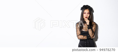 Thoughtful elegant evil witch in black lace dress, gothic wreath and makeup looking upper left corner. Woman making plans for halloween party, thinking over white background 94978270