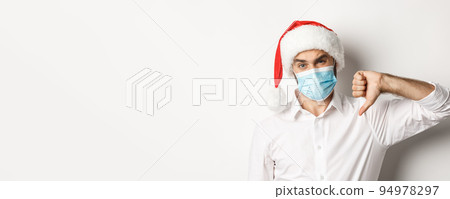 Concept of covid-19, social distancing and winter holidays. Close-up of disappointed man in face mask and christmas hat showing thumb down, standing over white background Concept of covid-19, social distancing and winter holidays. Close-up of disappointed man in face mask and christmas hat showing thumb down, standing over white background 94978297