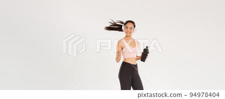 Full length of smiling attractive asian female athelte, girl doing fitness workout in gym, sportswoman or runner drinking water from bottle, listening music in wireless headphones, tossing hair 94978404