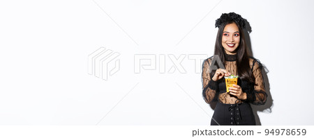 Smiling cute asian woman celebrating halloween, holding sweets and grinning happy, trick or treating in witch costume 94978659
