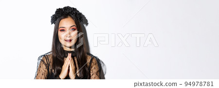 Close-up of young asian woman in black gothic dress and wreath holding hands in pray, girl wearing witch costume and celebrating halloween Close-up of young asian woman in black gothic dress and wreath holding hands in pray, girl wearing witch costume and celebrating halloween 94978781
