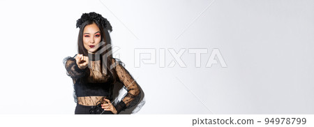 Confident beautiful asian woman in sexy witch costume lure you, tell come closer with extended finger, celebrating halloween and wearing gothic dress, standing over white background 94978799